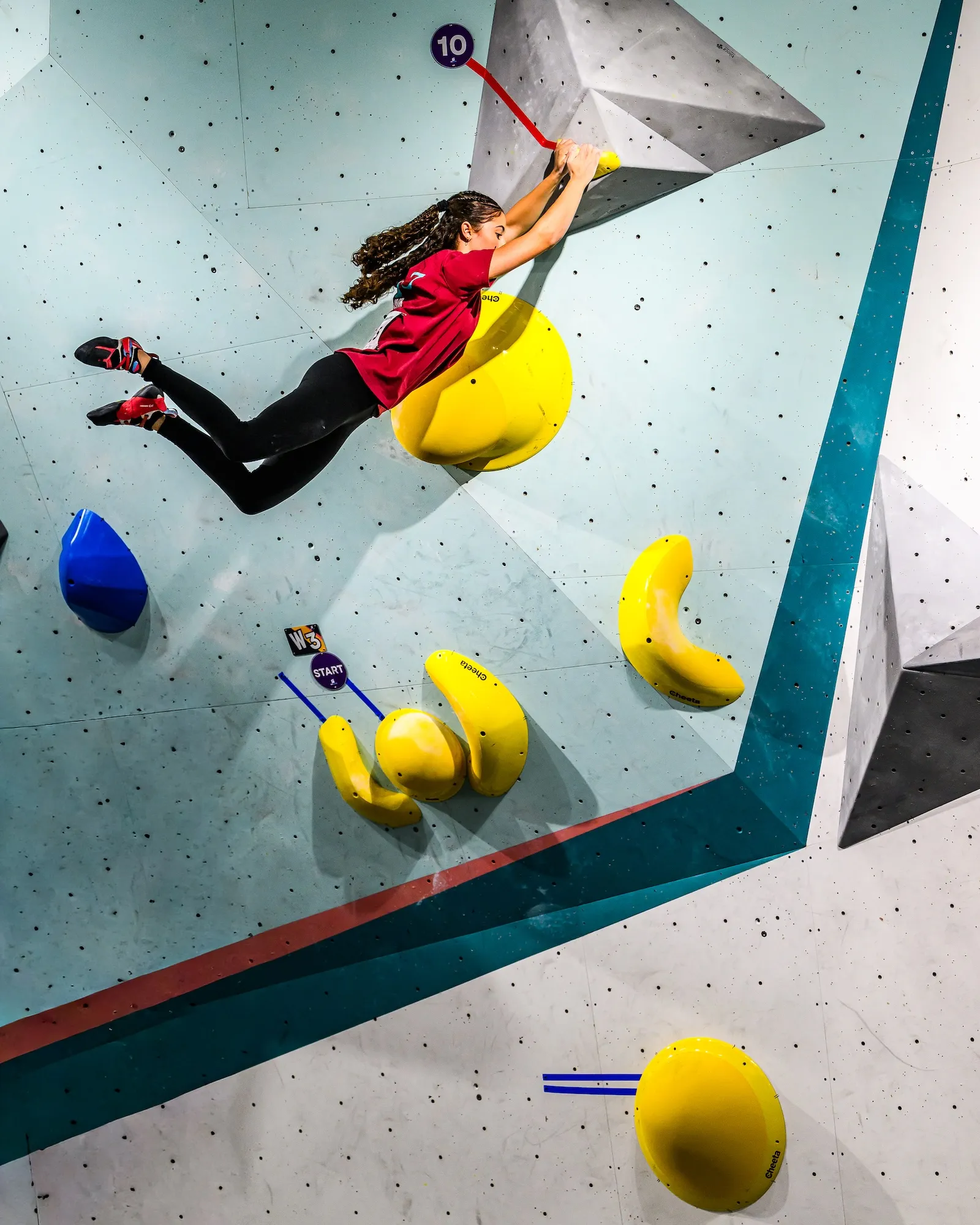 A climber swings almost horizontal off the teal wall, feet cut loose from the yellow volumes